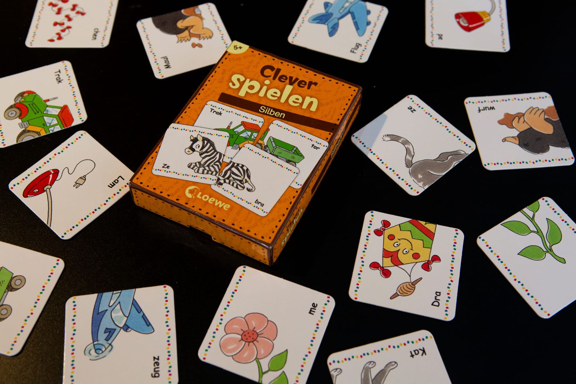 A colorful educational board game for children titled "Clever spielen Silben" (Clever Play Syllables) displayed on a black surface. The orange game box, marked with "5+" for ages 5 and up, is partially open, revealing cards inside. Around the box, numerous square cards with white backgrounds, colorful borders, and playful illustrations (e.g., a tractor, airplane, cat, flower, and kite) are scattered. Each card features a German syllable or partial word, designed to teach syllable recognition.