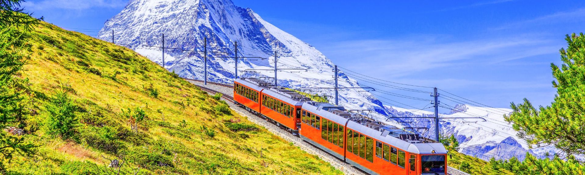 Panoramic view of a vibrant mountain landscape featuring an orange and silver passenger train on a curved track. The train ascends a grassy hillside with scattered trees, set against a backdrop of a towering, snow-capped mountain under a clear blue sky. Electrical poles and wires run alongside the track, emphasizing the engineering marvel within the natural beauty.