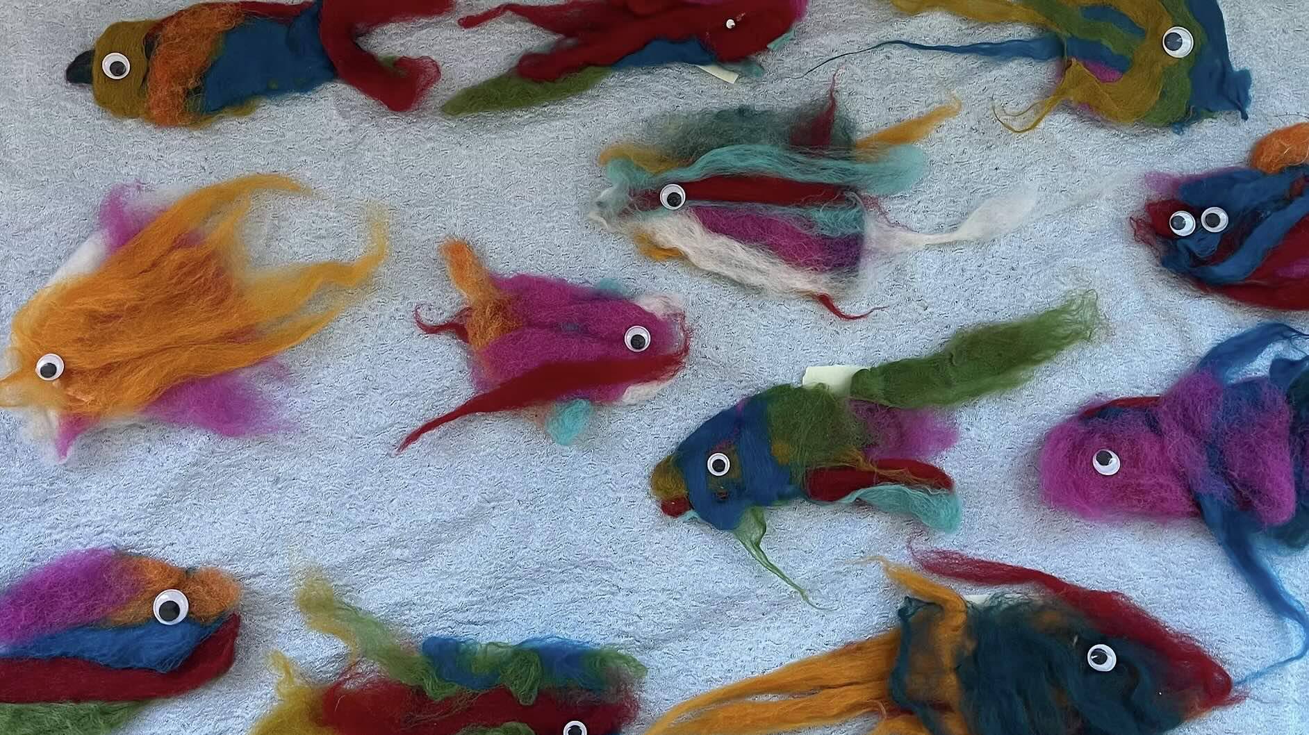 A collection of colorful, handmade felted wool fish displayed on a light blue textured surface. Each fish is uniquely designed with vibrant hues like orange, pink, blue, green, and yellow, featuring playful googly eyes and flowing fins. The fish vary slightly in size and shape, creating a whimsical and artistic arrangement.