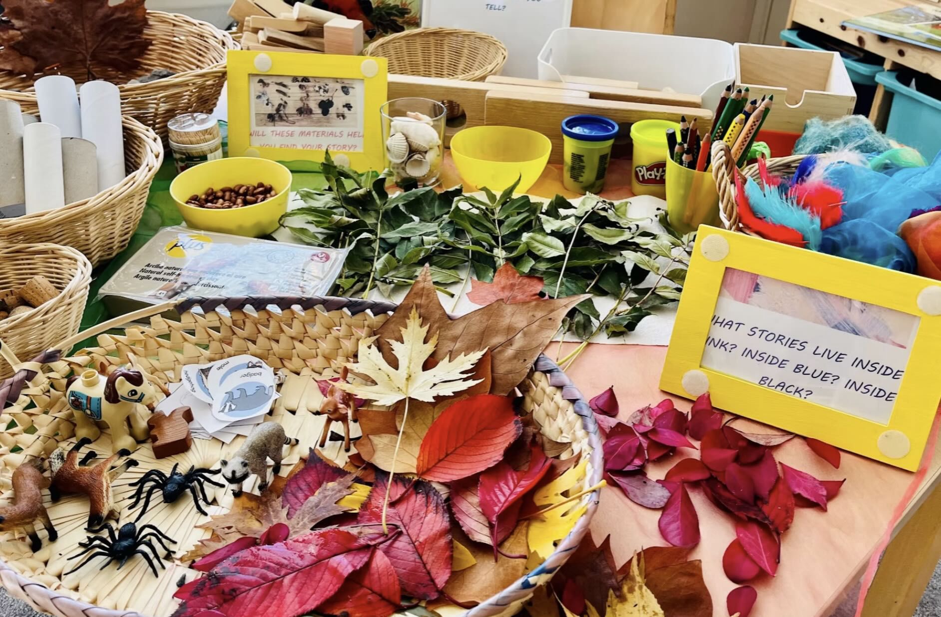 A creative play setup featuring a variety of natural and craft materials on a table. A woven basket holds colorful autumn leaves, small animal figurines, and white cards with animal names like 'hedgehog' and 'badger.' Surrounding the basket are green leafy branches, corks, cardboard tubes, and bowls with beans and seashells. Yellow frames display prompts like 'Will these materials help you find your story?' and 'What stories live inside pink, blue, black?' The scene encourages imaginative storytelling and hands-on exploration.