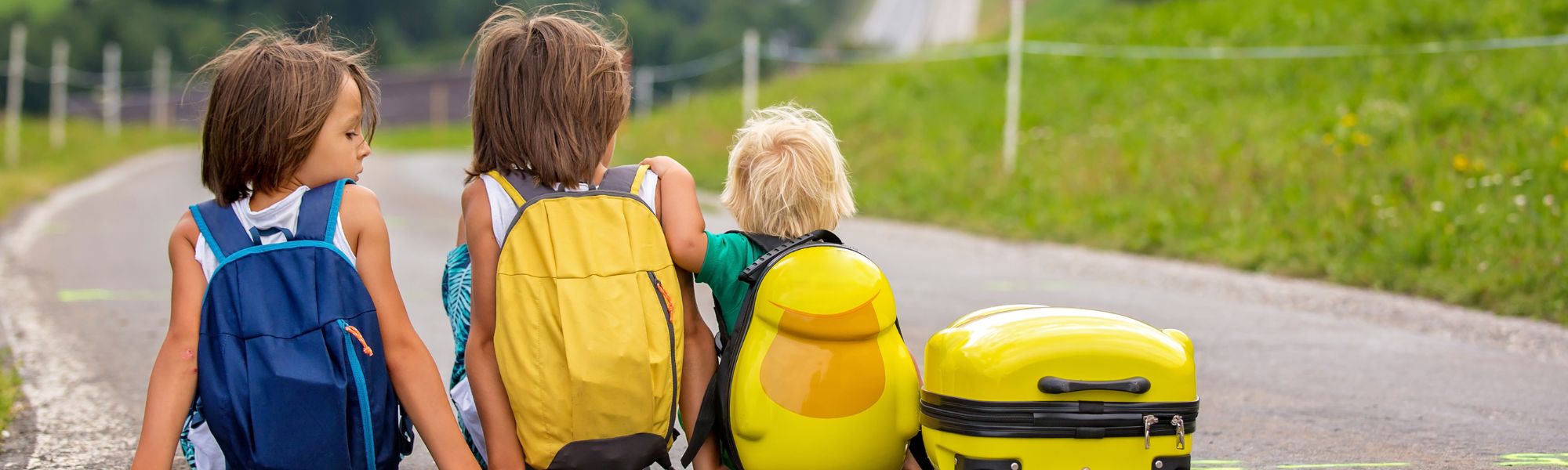 Three children with backpacks walk away from the camera on a paved path surrounded by green grass and trees. The child on the left wears a blue backpack, the middle child a yellow backpack, and the right child a bright yellow cartoon-themed backpack. A small yellow suitcase rests on the path beside them, suggesting a journey or adventure.