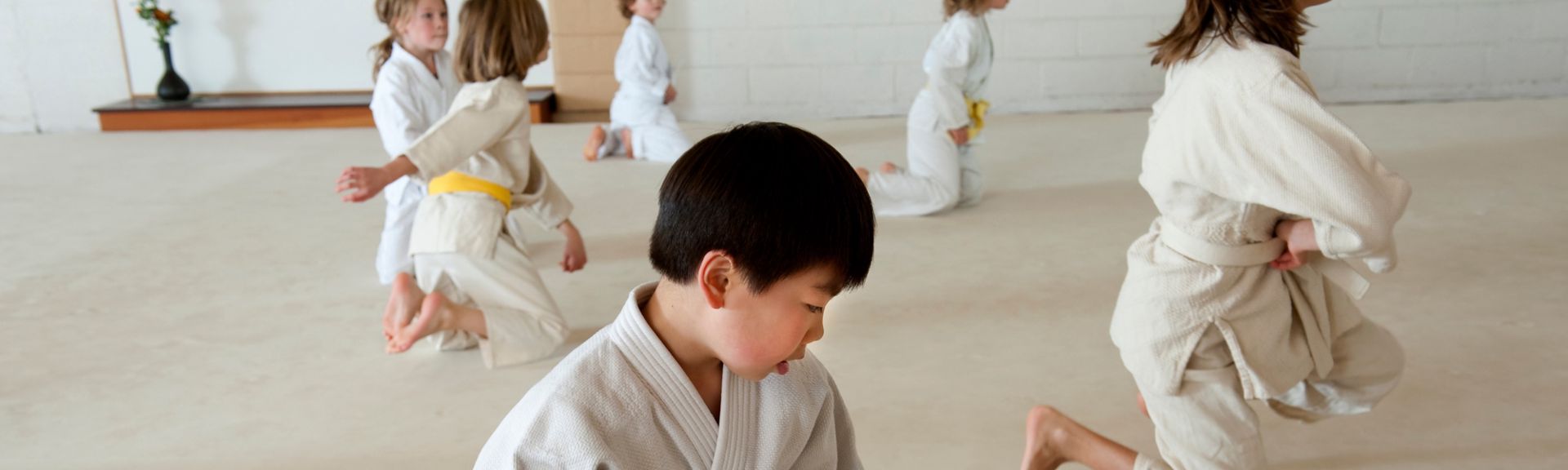 Group of children in white judo uniforms practicing on a training mat, with a young boy in the foreground focused on a kneeling stance, illustrating martial arts as a structured after-school activity for kids with ADHD.
