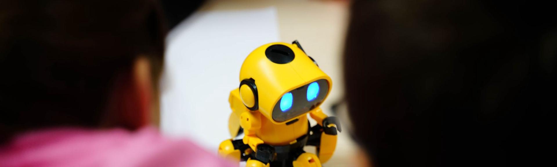 Close-up of a yellow and black toy robot with glowing blue eyes, positioned slightly off-center. The robot is surrounded by blurred figures of people in the foreground and mid-ground, suggesting an interactive or educational setting. The background is softly out of focus, emphasizing the robot as the main subject.