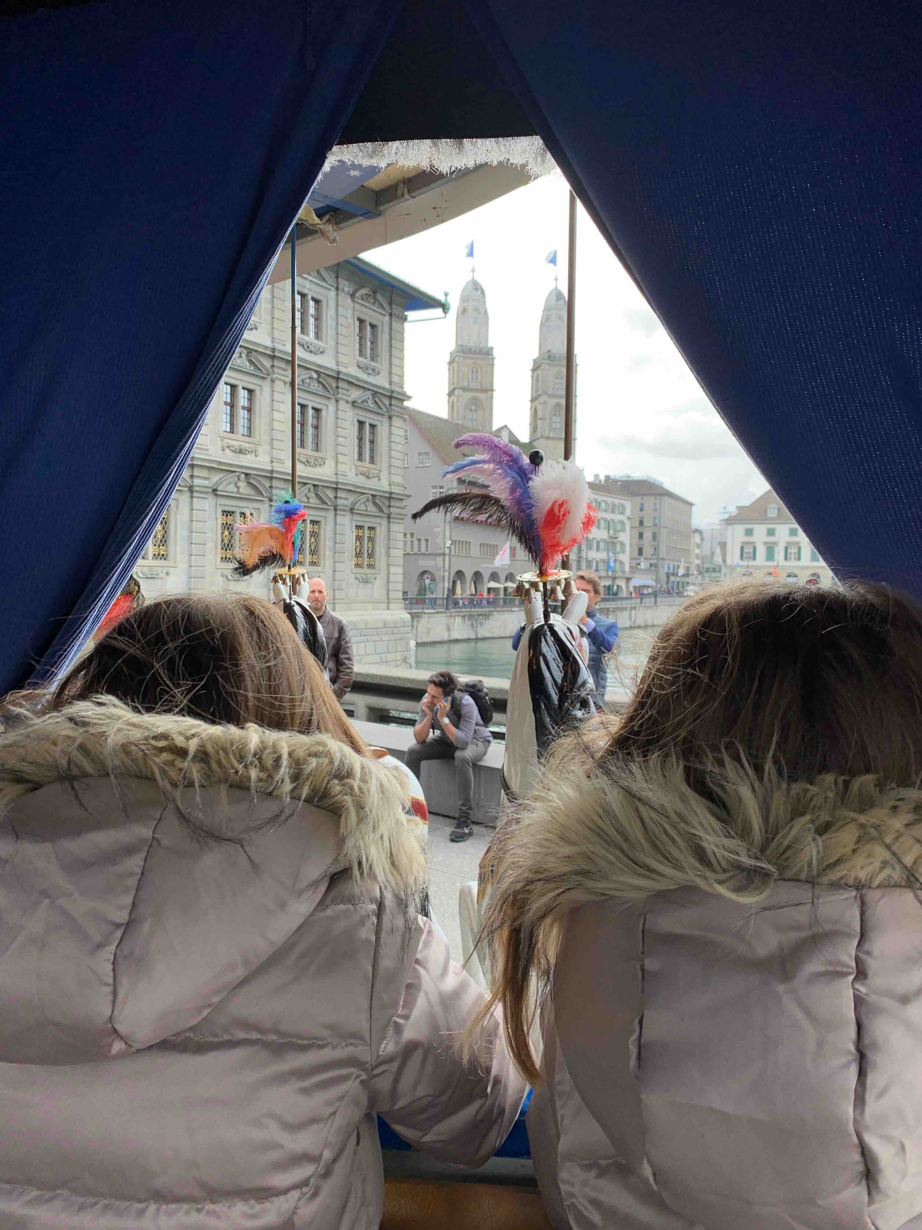 View from a horse-drawn carriage in Zurich's Old Town, framed by blue curtains, showing two passengers in fur-trimmed jackets, decorative plumes on the horse's harness, and the iconic twin towers of the Grossmünster church in the background.