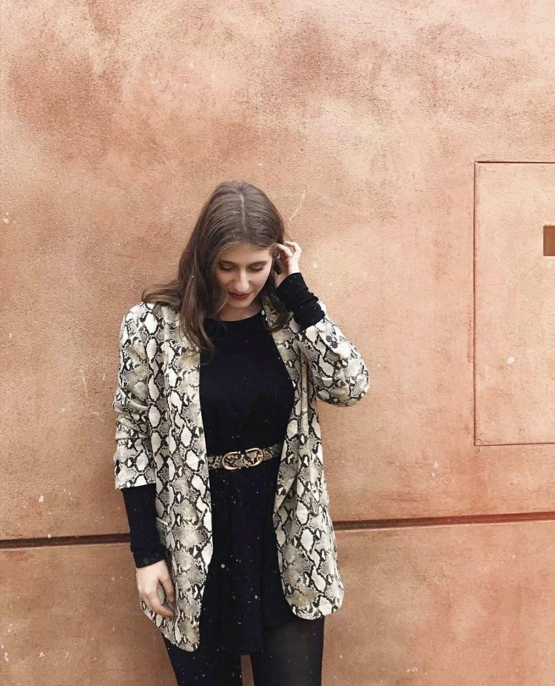 Νεαρή γυναίκα με μαύρο φόρεμα και σακάκι σε snake print στέκεται μπροστά σε τοίχο σε απόχρωση τερακότας, κοιτάζει προς τα κάτω και αγγίζει τα μαλλιά της.