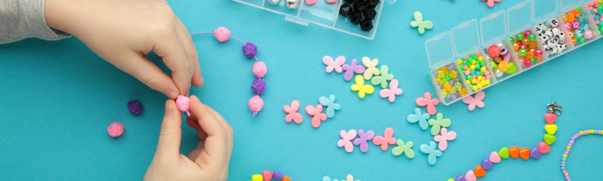 Flat lay of a child's hands crafting beaded jewelry on a vibrant teal surface. The child threads a pink spherical bead onto a purple string, surrounded by colorful butterfly-shaped beads in pastel shades. Two clear organizer boxes hold an assortment of beads, including silver, black, faceted, and lettered beads, as well as translucent and opaque colorful beads. Partially completed bracelets with heart-shaped and round beads are visible in the bottom right corner, showcasing a creative and vibrant jewelry-making activity.