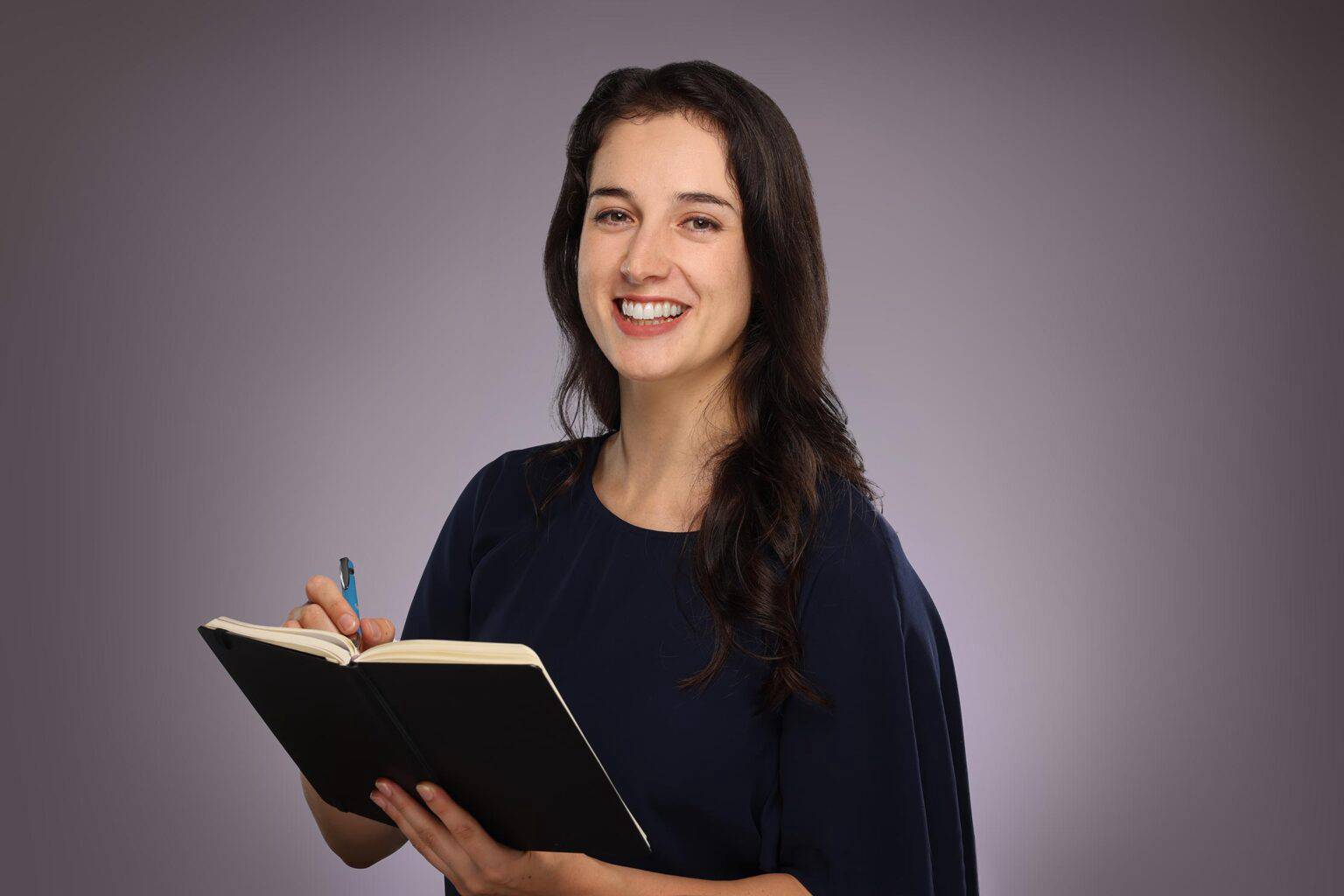 Smiling woman holding notebook and pen wearing dark blue shirt with grey background