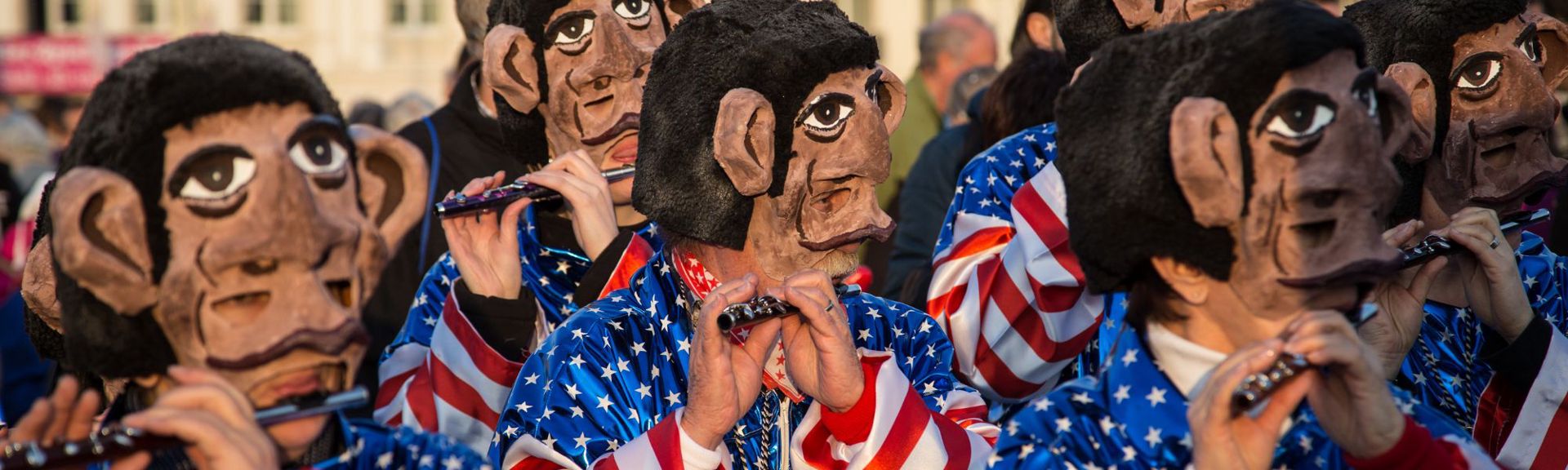 Group of performers wearing brown monkey-like masks with exaggerated features and black fuzzy hair, dressed in American flag-themed outfits, playing flutes during an outdoor event or parade.