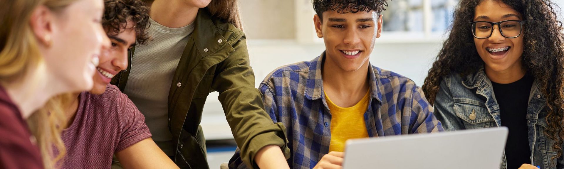 A group of smiling teenagers gathered around a laptop, collaborating on a school project and learning together in a classroom setting using AI constructively. 