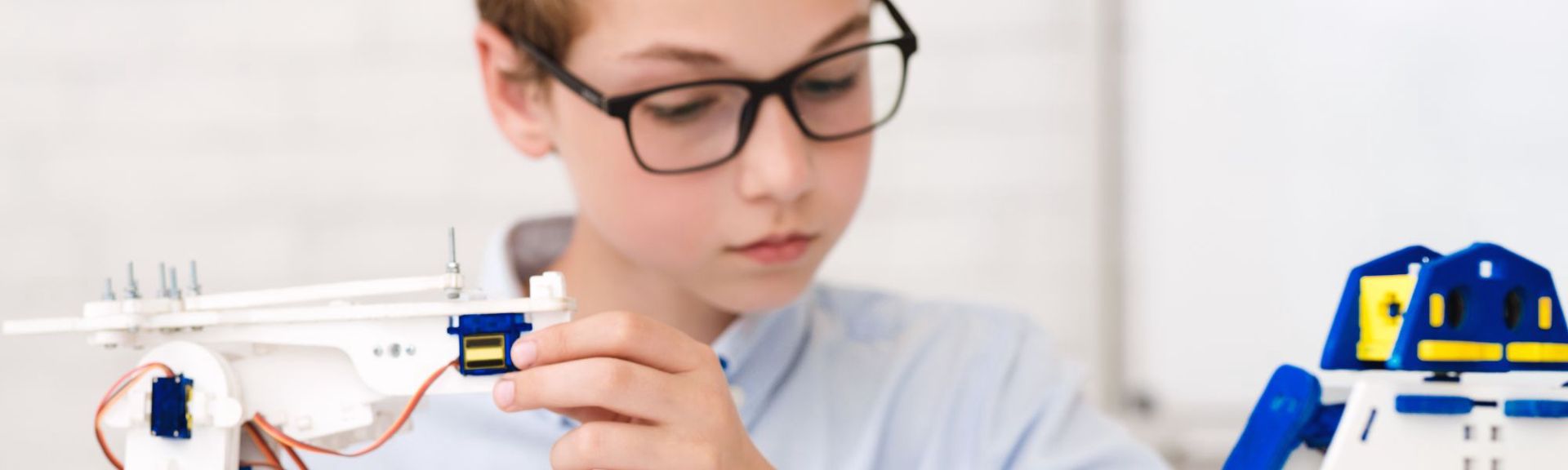 A young boy, wearing black-rimmed glasses and a light blue collared shirt, is focused on assembling a white robotic or electronic device. He holds a component with visible screws and a blue and yellow servo motor, connected by orange and brown wires. To his right, a partially visible blue and white robot with yellow accents adds context to the STEM activity. The background is softly blurred, suggesting an indoor educational setting like a classroom or workshop. The scene highlights hands-on learning and engagement with robotics and technology.