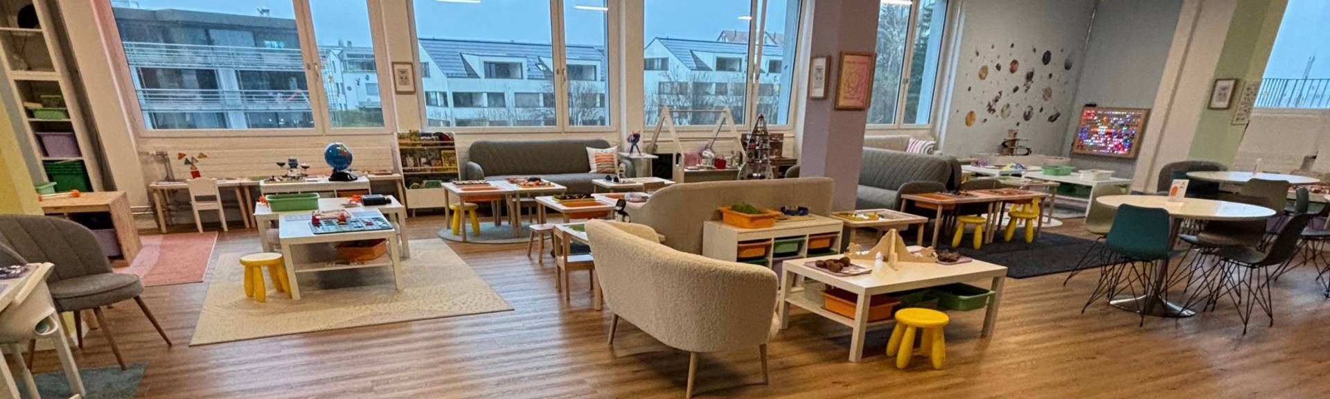Wide view of EduPlay STEM Lab's open-play room in Steinhausen, with low activity tables, a bookshelf, soft armchairs and sofas, and floor-to-ceiling windows overlooking the neighborhood.