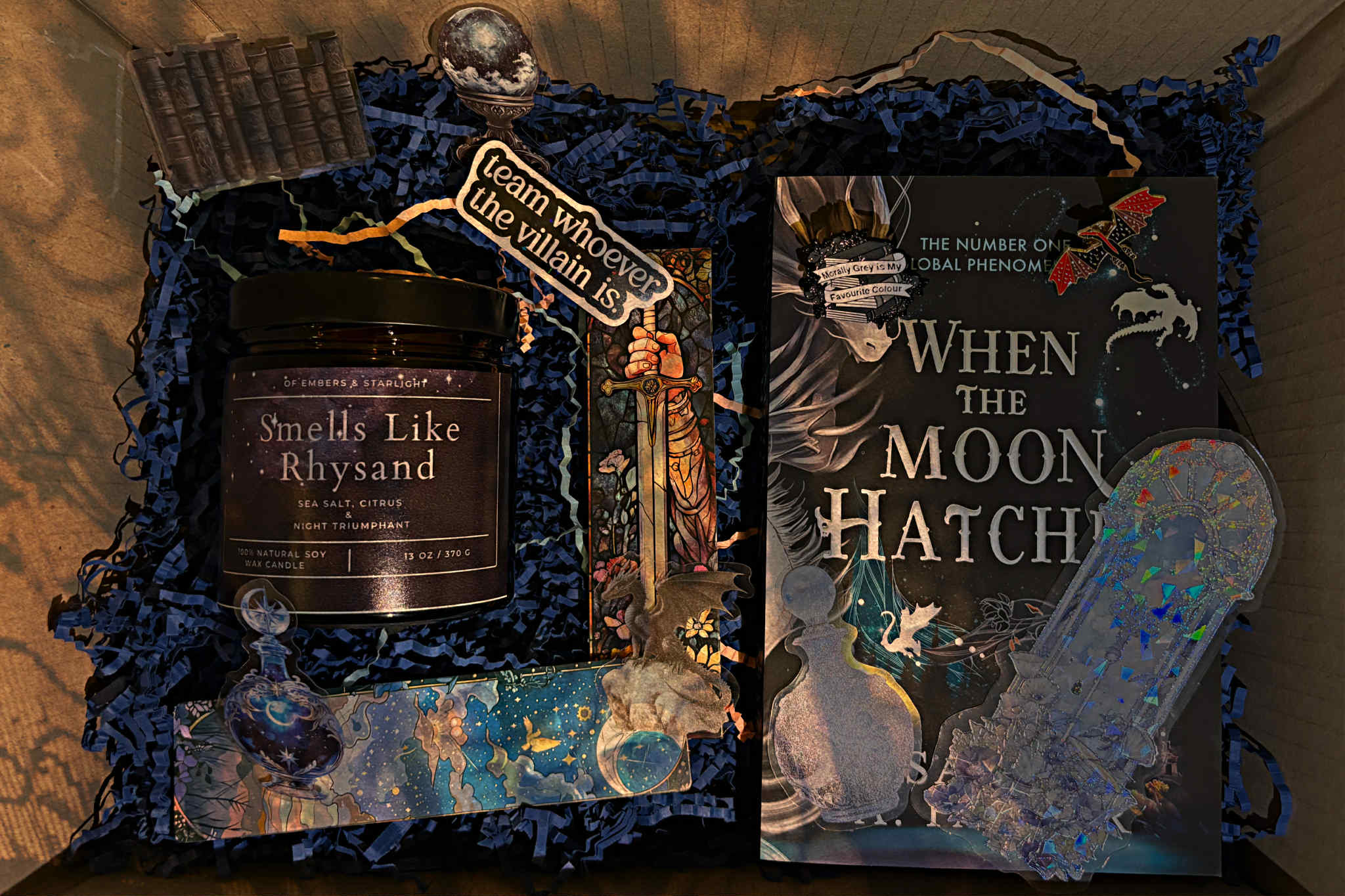 Book-themed gift box with a “Smells Like Rhysand” soy candle, the fantasy novel When the Moon Hatches, and assorted decorative items including dragon and potion stickers, a sword illustration, and holographic fantasy bookmarks arranged on dark shredded paper.