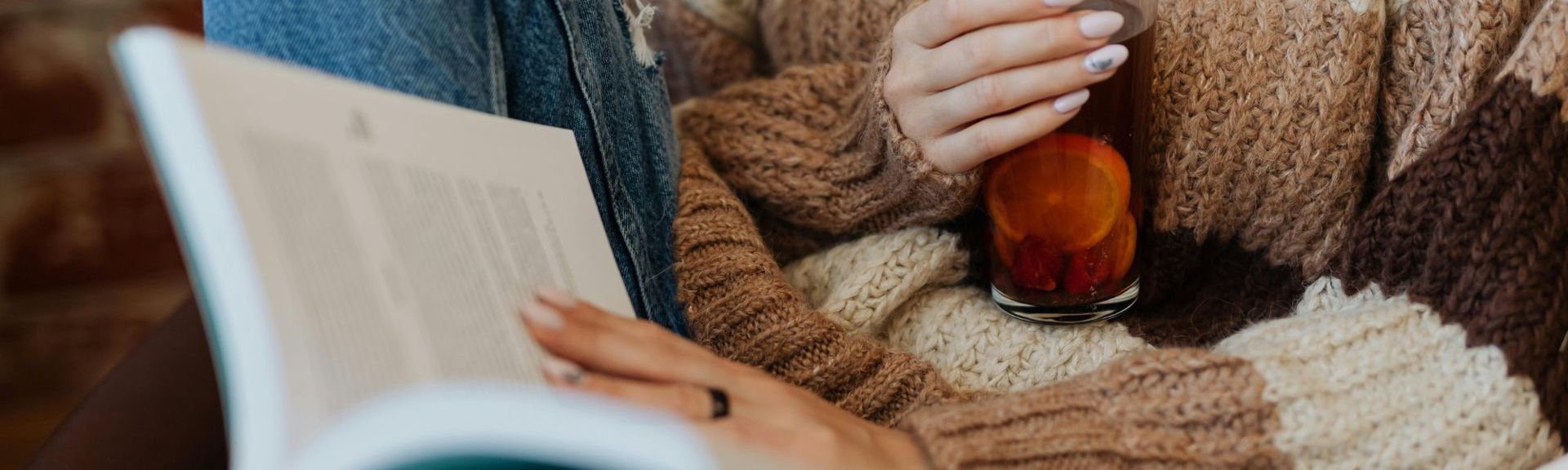 Close-up of a person in a chunky knit sweater and jeans relaxing with an open book on their lap, holding a glass of hot fruit tea with orange slices and berries.