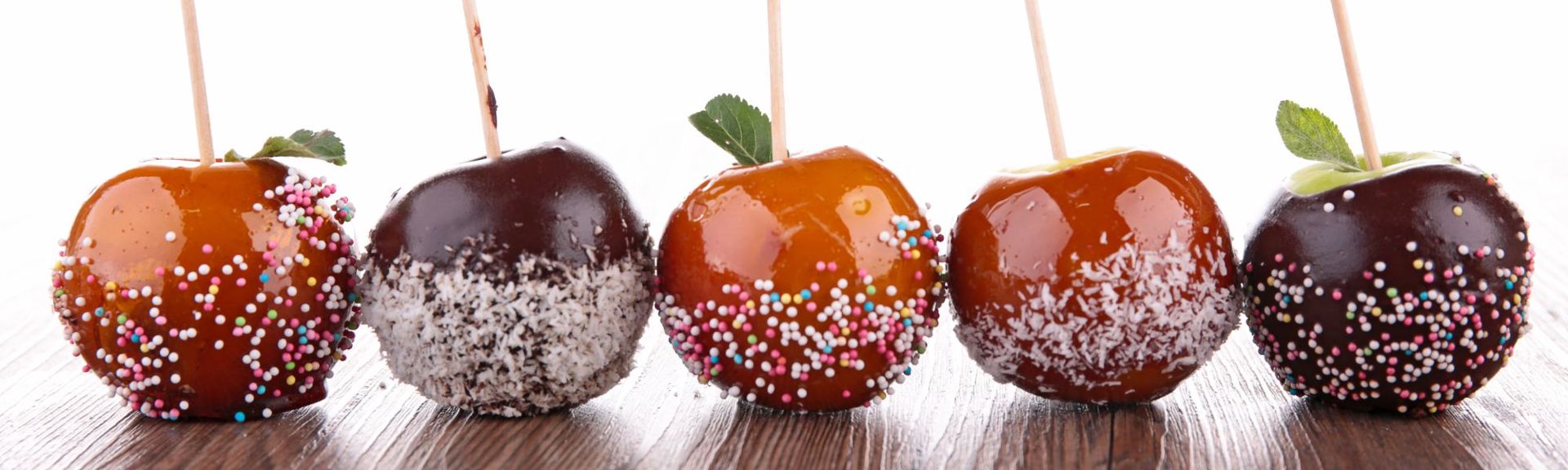 Alt Text: A row of five candy apples on a wooden surface against a white background. The apples alternate between caramel and chocolate coatings, with toppings of colorful sprinkles or shredded coconut. Each apple has a wooden stick and some feature small green leaves at the base.
