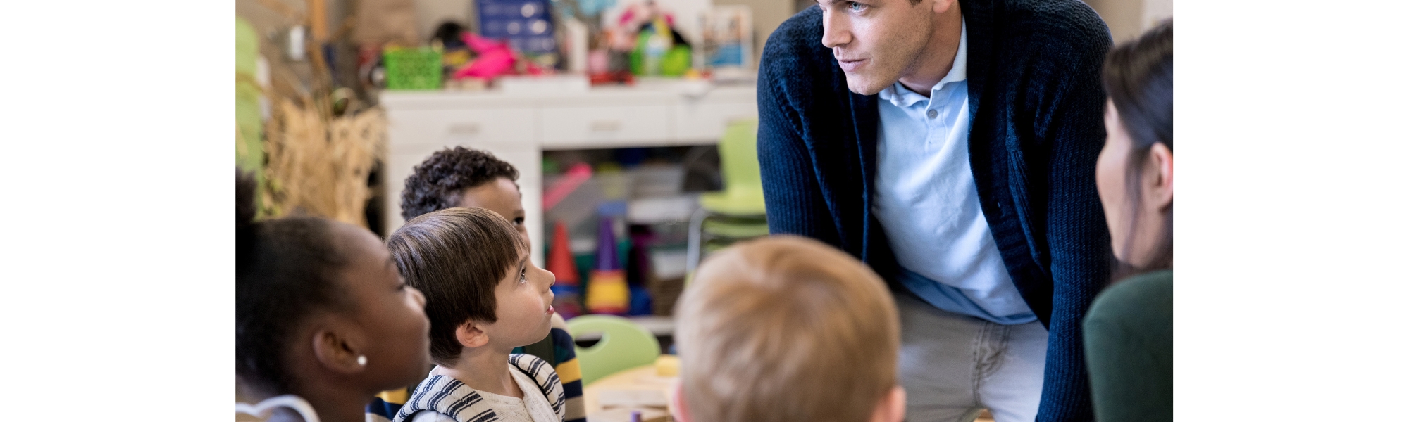 Teacher kneeling to talk with a group of young students in a classroom