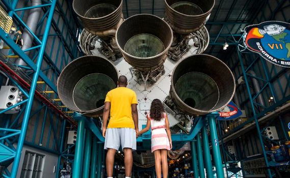 An adult and a child stand hand-in-hand, gazing up at the colossal engines of a rocket on display in a museum. The massive nozzles dominate the scene, with mission patches and structural beams in the background, emphasizing the scale and history of space exploration.