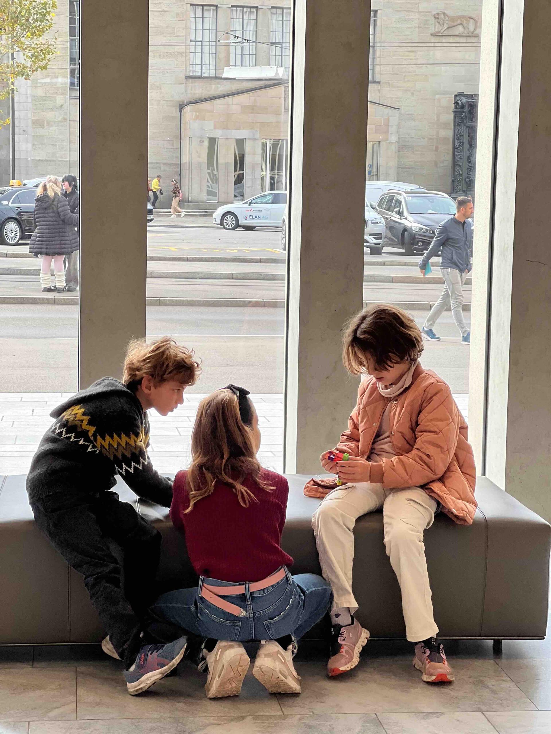 Three children play indoors on a grey bench near floor-to-ceiling windows, with a bustling Zurich street scene visible outside, including pedestrians, cars, and a classical stone building.