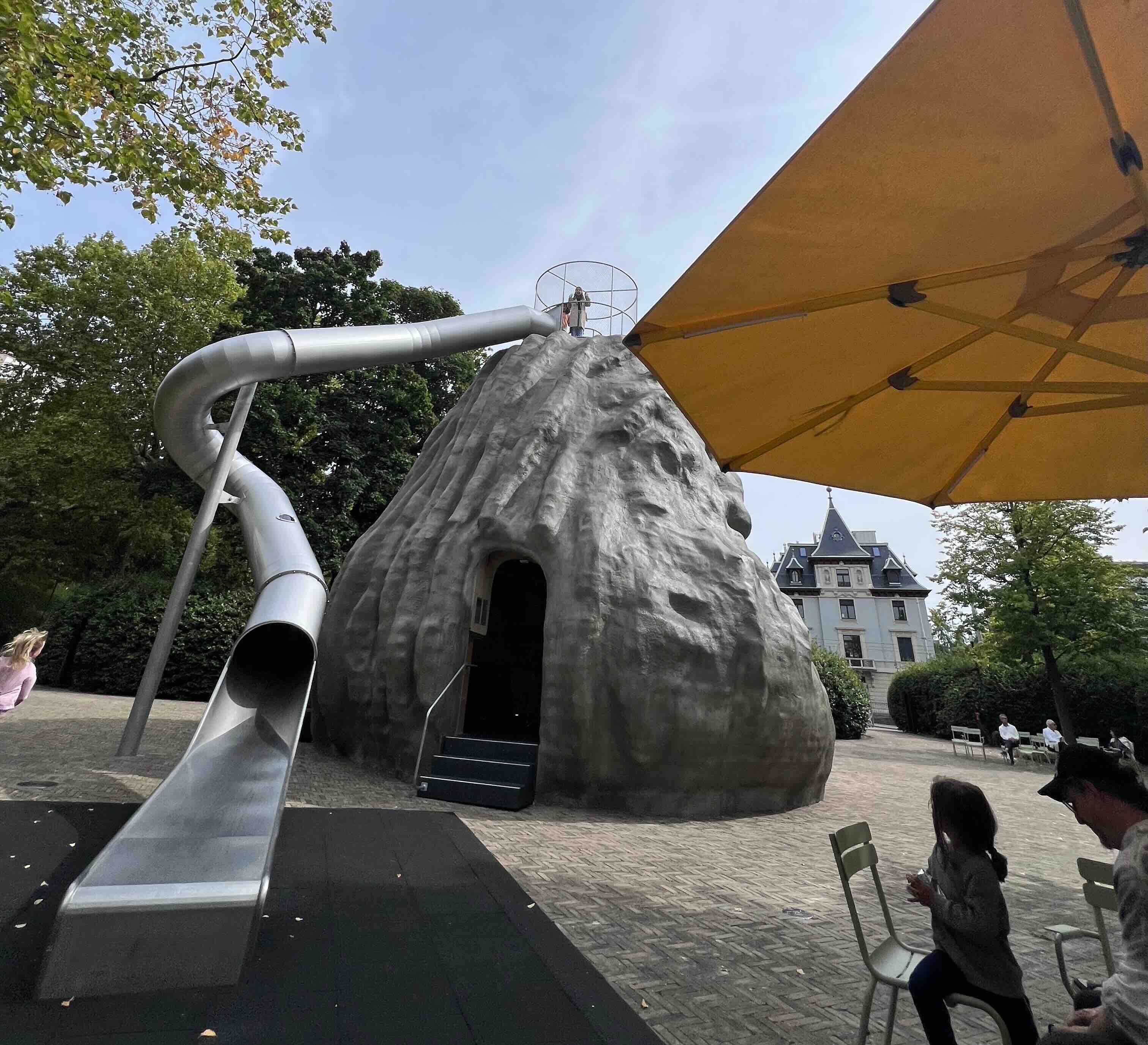 A large grey climbing structure with a gleaming silver slide set in a park-like environment, surrounded by leafy trees, a yellow umbrella, and a stately European-style building in the background.