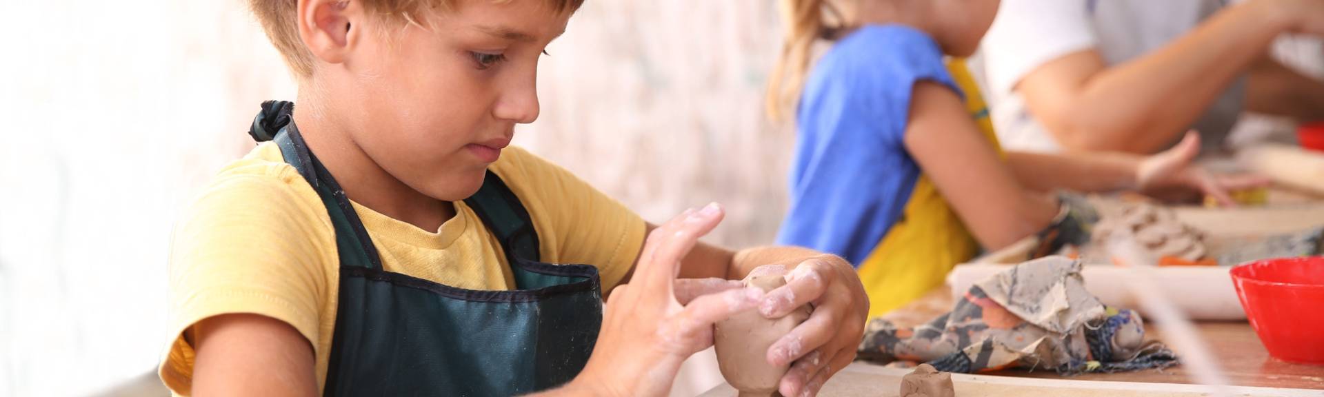 Close-up of a boy in a green apron shaping wet clay by hand at LOKI Studio's pottery table, with a hand guiding him and other kids working with clay in the background.