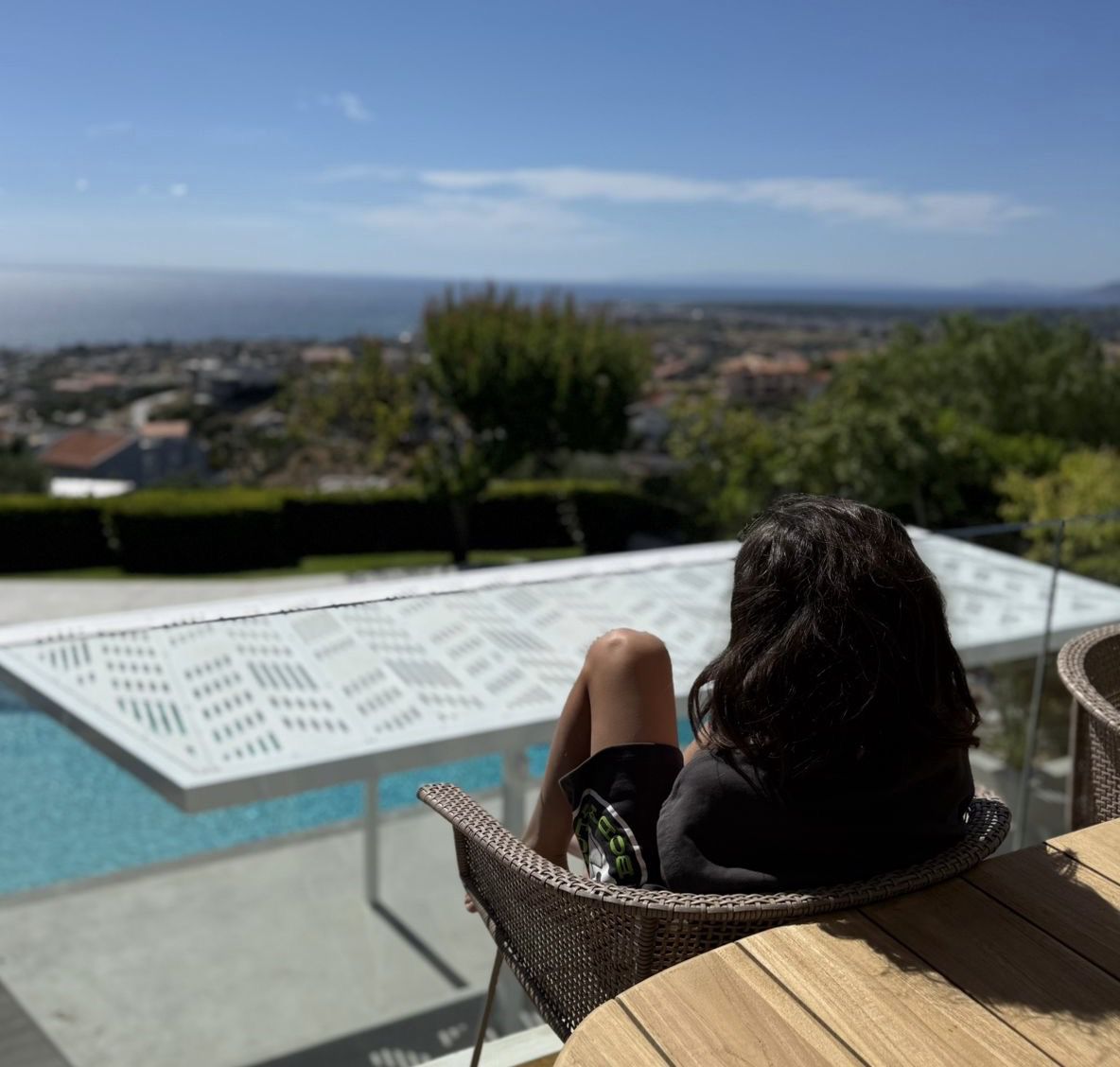 A young boy with long hair looking over a pool into the sea in the distance in Rafina, Greece
