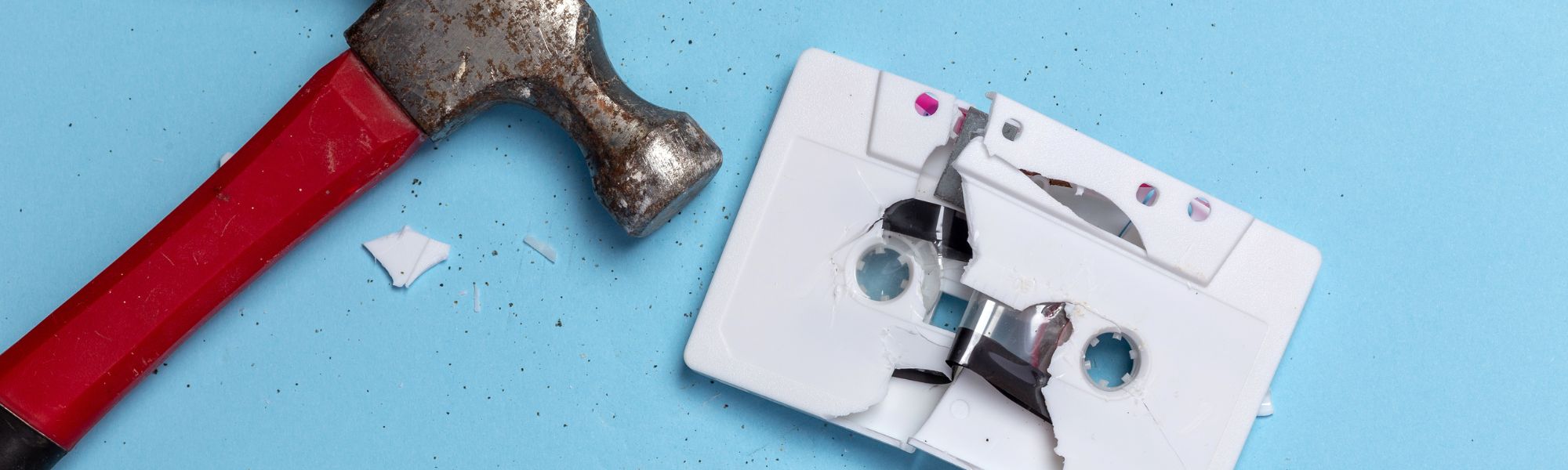 Red-handled hammer beside a smashed white cassette tape on a light blue background.