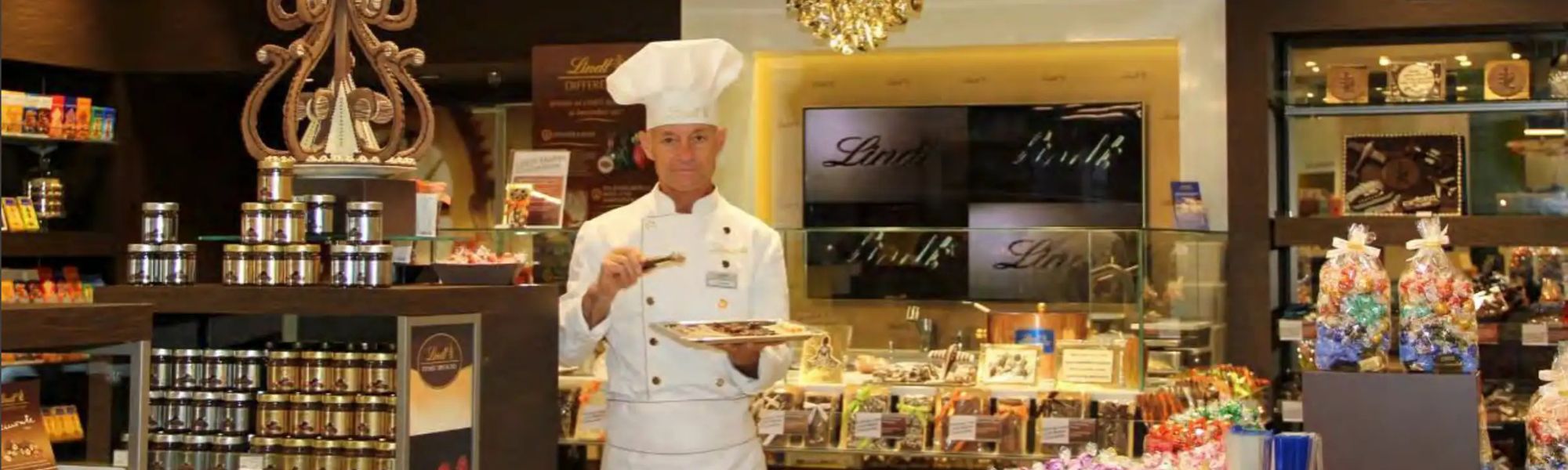 A Lindt Maître Chocolatier stands in a premium chocolate shop, wearing a white chef's uniform and hat, holding a tray of chocolates in one hand and a chocolate-dipping tool in the other. Behind him, shelves are filled with colorful chocolate products, including jars, gift-wrapped assortments, and individually wrapped truffles. A large display screen prominently features the Lindt logo, while the warm lighting and elegant decor create an inviting atmosphere.