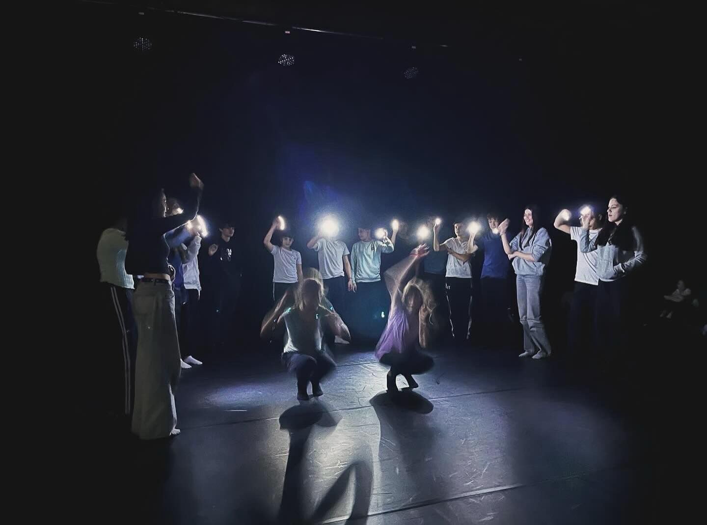 A group of dancers on a stage standing around in a semi-circle around two dancers that are performing a dancing duet that looks like breakdancing.  The surroundings are black with the only light coming from the mobile phones of the crowd watching the two dancers in the center. Their face is blurry.