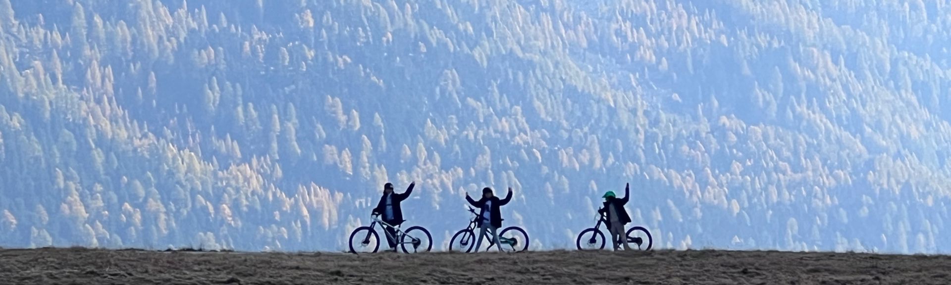 Three kids on mountain bikes standing on a ridge with their bikes, set against a steep, forested mountainside. The bikers raise their arms in a celebratory gesture, surrounded by earthy terrain and autumn-colored trees under a hazy, natural light.