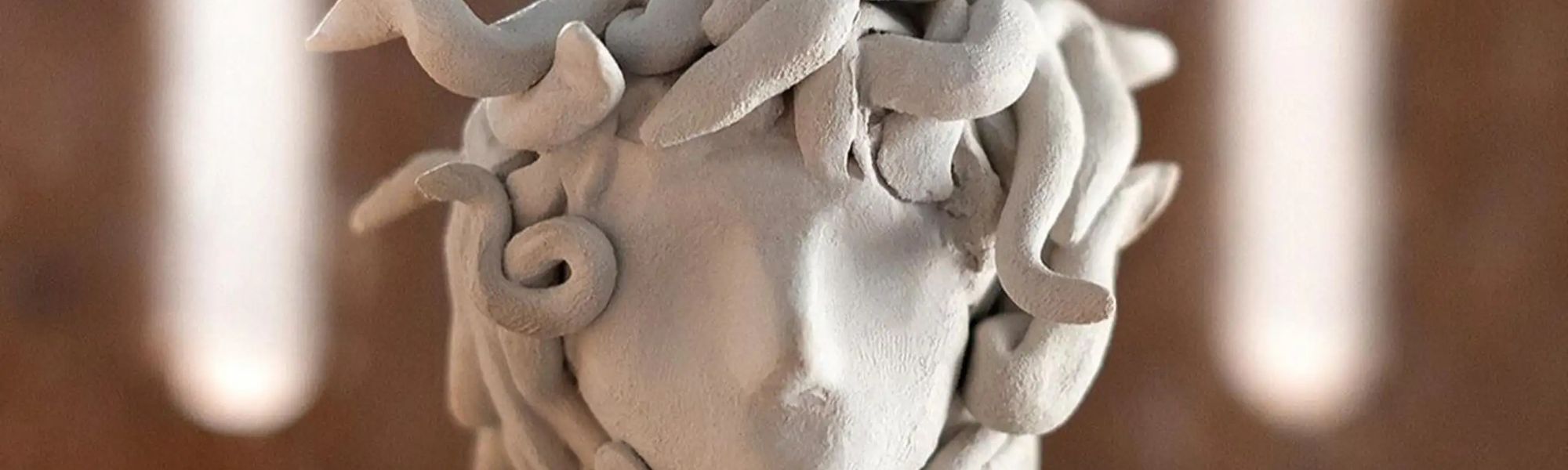 Head-on view of a light-colored clay sculpture depicting a stylized head with snake-like forms as hair, set against a warm brown background with soft lighting.