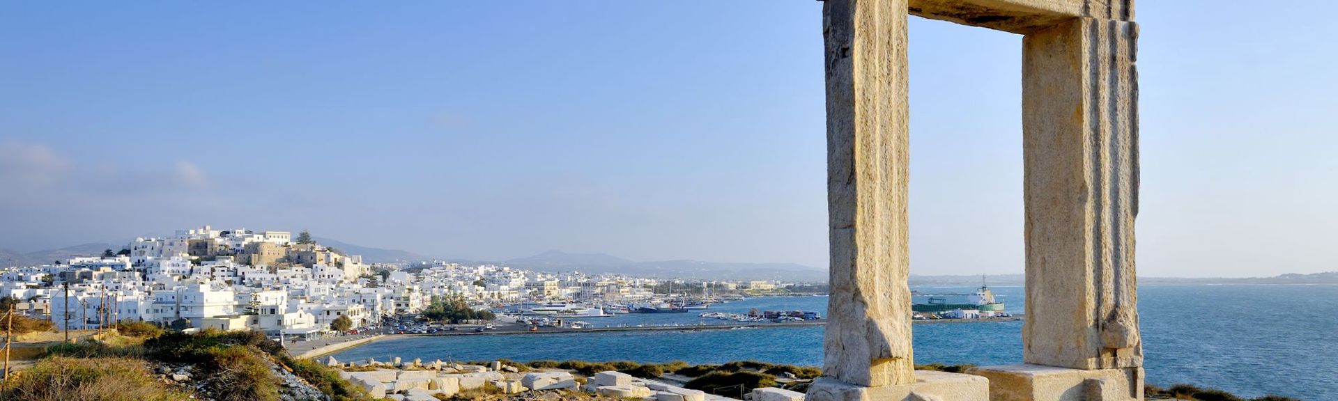 Panoramic view of Naxos, Greece, featuring the ancient Portara, a large stone doorway from the unfinished Temple of Apollo, in the foreground. The Portara frames a coastal scene with a whitewashed Cycladic town (Chora) on a hillside, a harbor filled with boats and a ferry, and rolling blue-gray hills in the background. The bright blue sea and sky contrast with the earthy tones of the rocky, grassy hill and the weathered stone pillars, highlighting the blend of ancient history and modern Mediterranean charm.