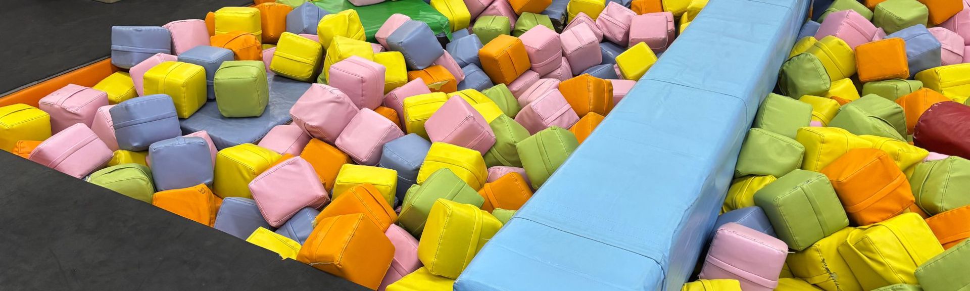 Colorful indoor playground with padded walls and soft play equipment, featuring cheerful cartoon decorations, flowers, animals, and interactive elements designed for young children.