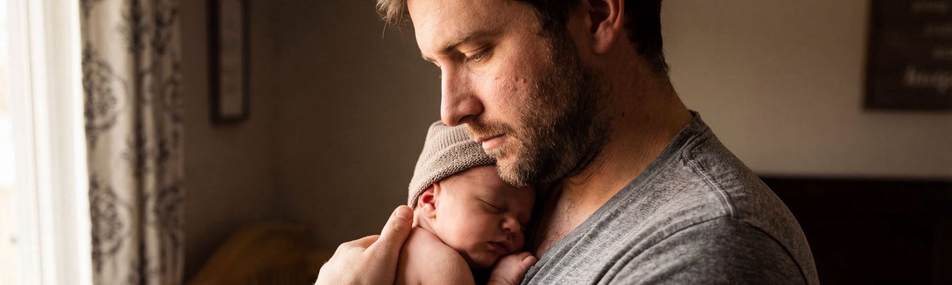 A serene portrait of a father holding his sleeping newborn baby against his chest. The father, wearing a grey v-neck t-shirt, gazes down at the baby, who is wearing a light brown knit beanie. Soft natural light highlights their profiles, creating an intimate and warm moment, with a blurred indoor background featuring muted tones and subtle decor.