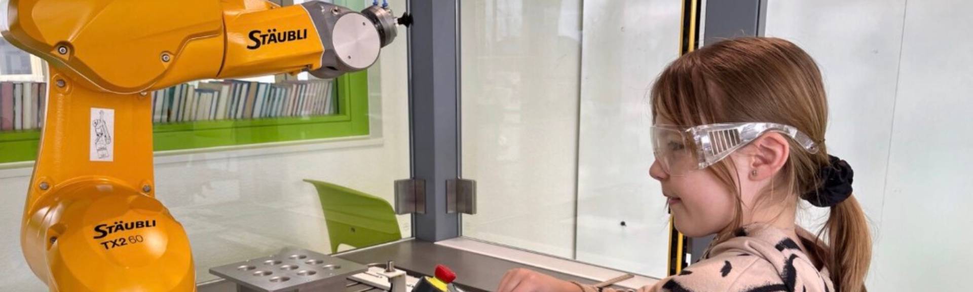 A student wearing safety glasses operates a Stäubli industrial robotic arm using a control pendant at OBS Wollerau's STEAM lab.