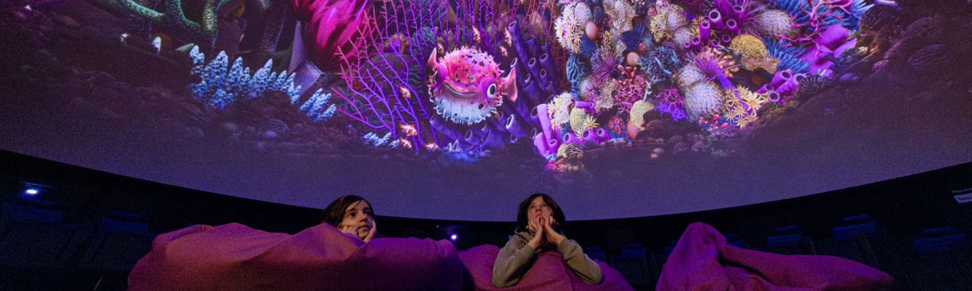 Children lying on beanbags inside a dome theatre in Luzern Verkehrhaus, watching a colorful underwater ocean animation projected on the ceiling by PROJEKTIL, called pixel zoo.