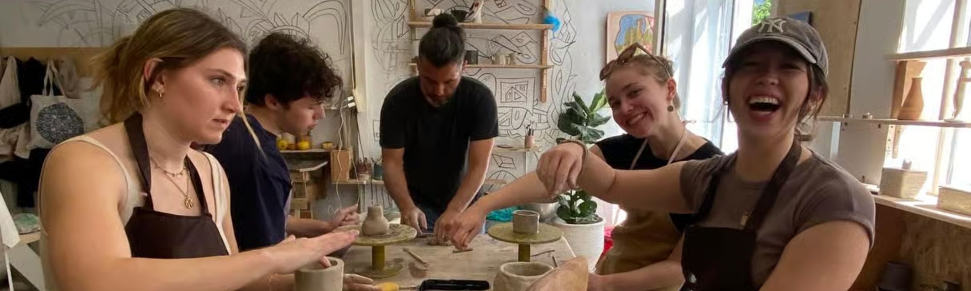 Five young adults in a pottery studio, actively shaping clay on a long table, with tools and unfinished pottery pieces scattered around. The scene is bright and filled with laughter and focus.