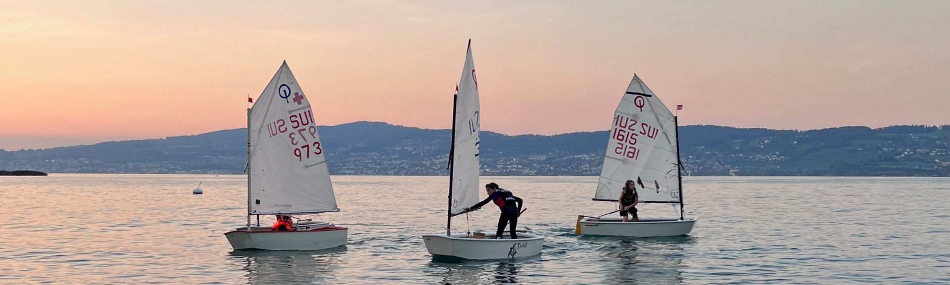 Three Optimist dinghies with young sailors on a calm Swiss lake during sunrise or sunset. The boats feature white sails marked with 'SUI' and numbers, indicating Switzerland. The serene scene includes rippling water, a distant lakeside town, rolling green hills, and a soft gradient sky transitioning from orange to lavender.