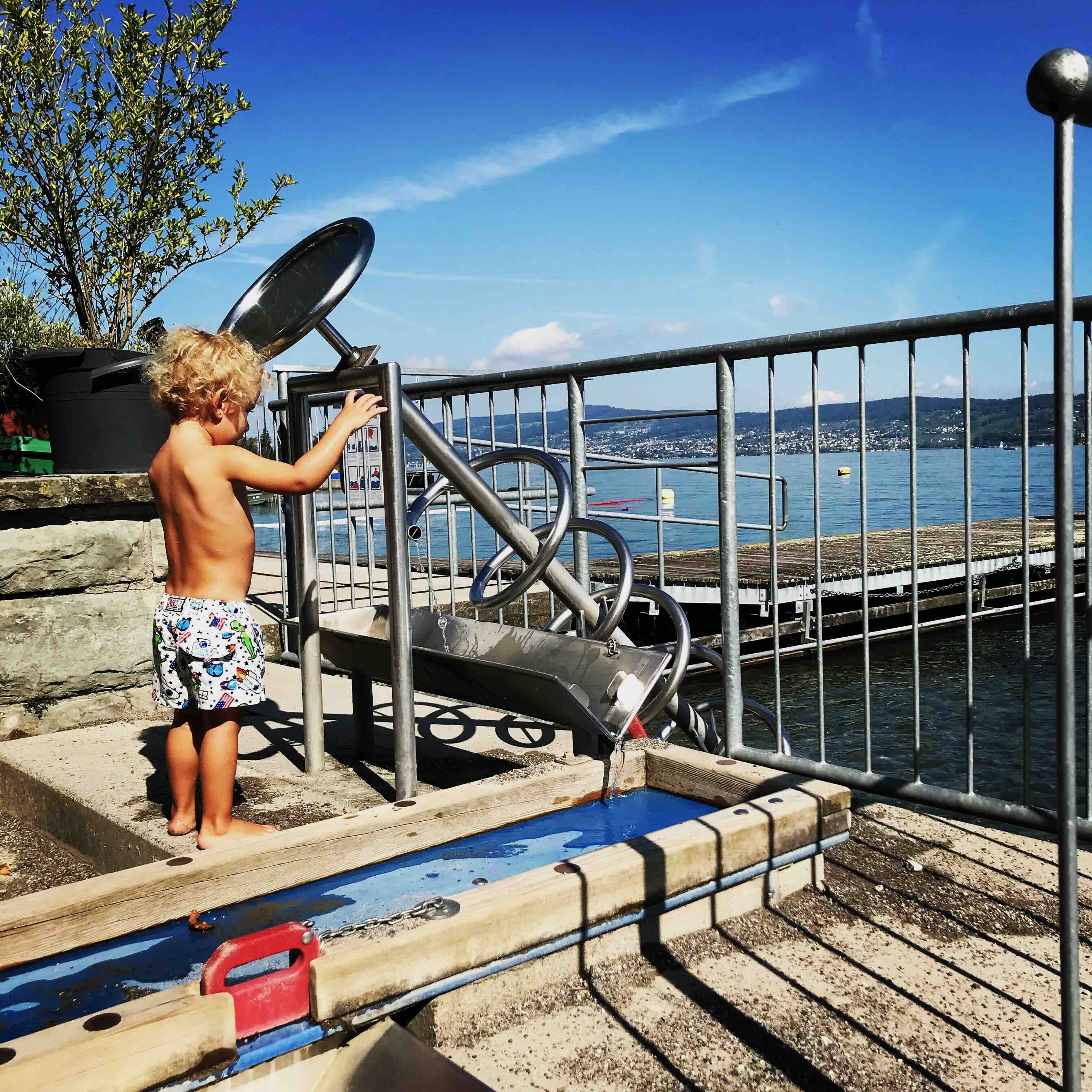 A young boy in blue swim trunks interacts with a stainless steel water play structure featuring an Archimedes' screw and flowing water, set against the backdrop of a serene lake and distant green hills under a clear sky.