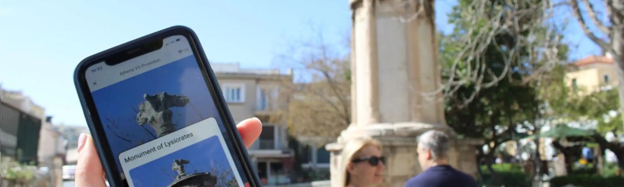 A person holding a smartphone showing an app with the title 'Athena VS Poseidon' and an image of the Monument of Lysicrates. The monument is visible in the sunny outdoor background, along with trees and other visitors.
