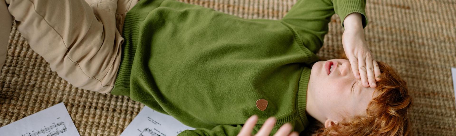 A young child with reddish-blonde curly hair lies on a textured light brown rug, dressed in a green long-sleeved top with a small circular patch on the chest and beige trousers. The child rests with one hand on their forehead, eyes closed, and a white sheet of music with visible notes lies nearby, suggesting a moment of rest or reflection during a musical activity.