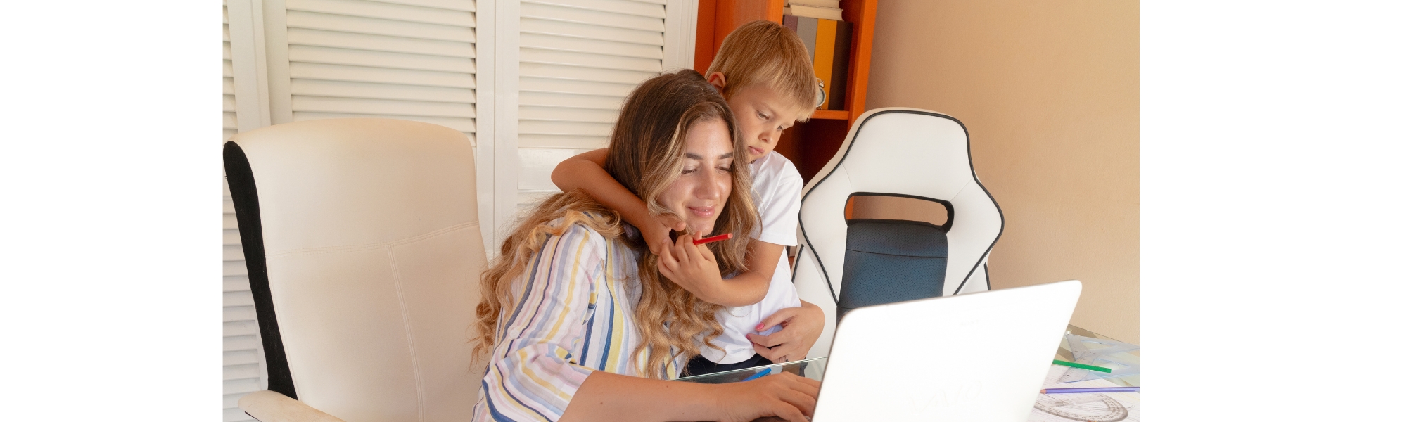 Eine lächelnde Mutter arbeitet zu Hause am Laptop, während ihr kleiner Sohn sie von hinten umarmt und einen roten Stift hält.