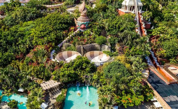 An aerial view of a tropical-themed water park surrounded by lush green foliage. The park features two large bowl water slides, a multi-level slide with a pagoda-like structure, turquoise leisure pools with people enjoying the water, and ornate statues with Southeast Asian-inspired designs. The bright sunlight highlights the vibrant greenery and colorful attractions, creating a lively and inviting atmosphere.