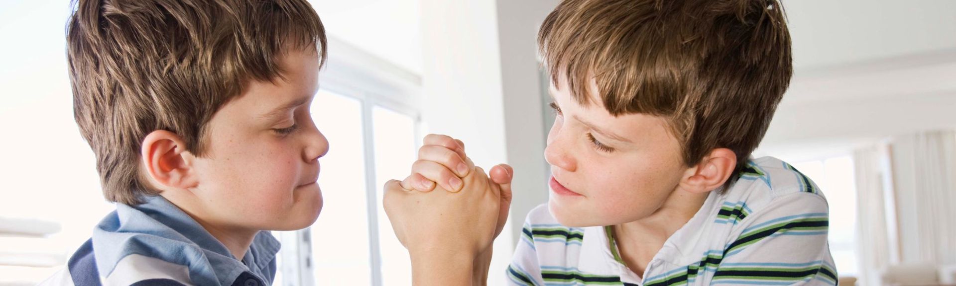 Two young boys, likely siblings or friends, engaged in a playful arm-wrestling contest. They sit face-to-face with their hands clasped in the center, both focused and concentrating. The background is softly lit, with bright, diffused light coming from windows, creating a warm and airy atmosphere.