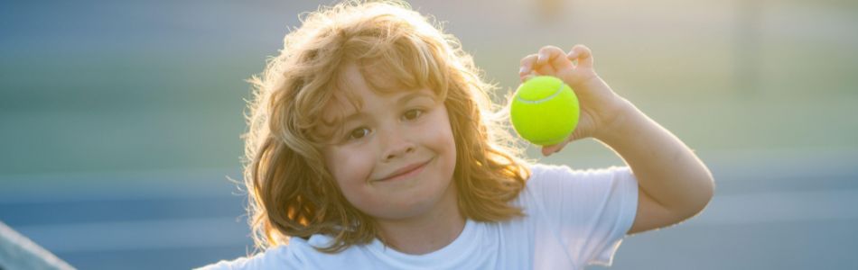 A cheerful young child smiles while holding up a tennis ball on a sunny outdoor court. The image captures the excitement of kids’ summer camps, where children stay active, build new skills, make friends, and enjoy memorable outdoor adventures.