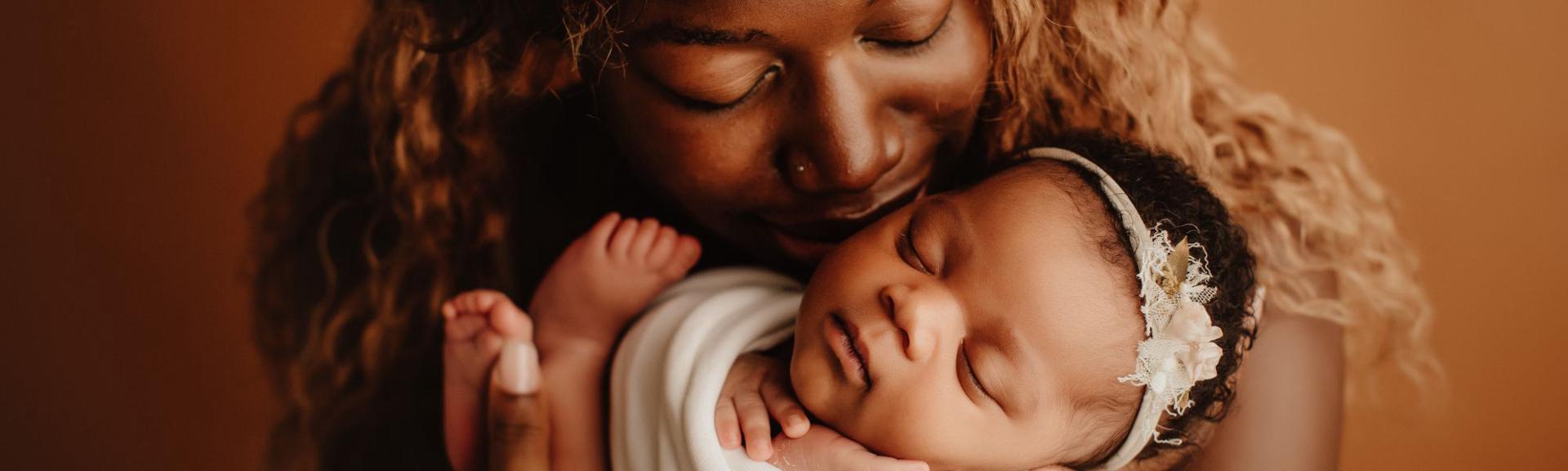 A tender moment between a mother and her newborn baby. The mother, with dark brown skin and curly auburn hair, holds her swaddled baby close. Both have their eyes closed, exuding peace and connection. The baby, wrapped in a soft white blanket, wears a delicate headband with floral embellishments. The warm, soft lighting highlights their bond against a muted terracotta background