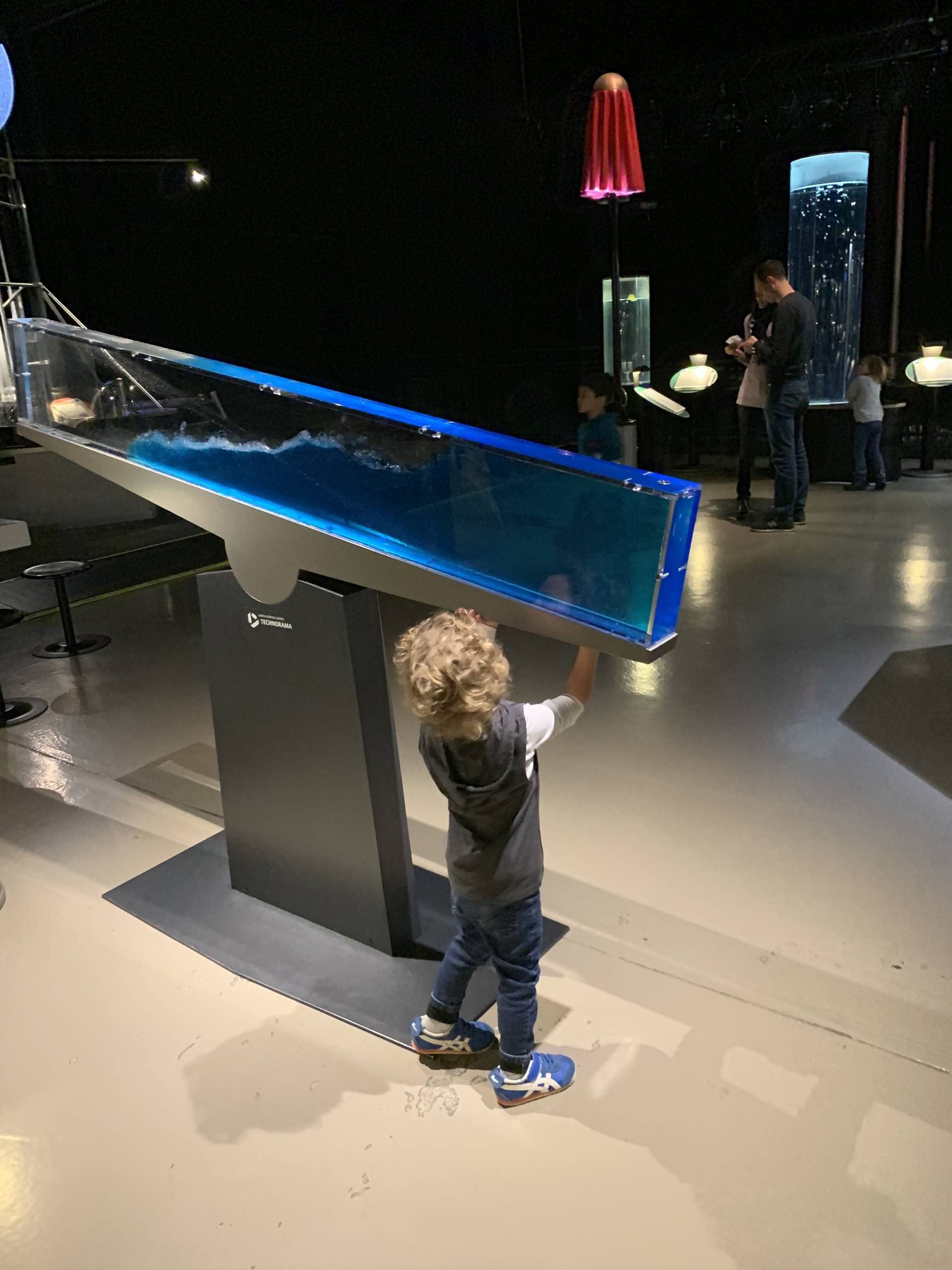 A young blond boy between 3 and 4 years old is observing the effects of water movement and sand along a rectangular aquarium that moves at the Swiss Science Center Technorama in Winterthur.  