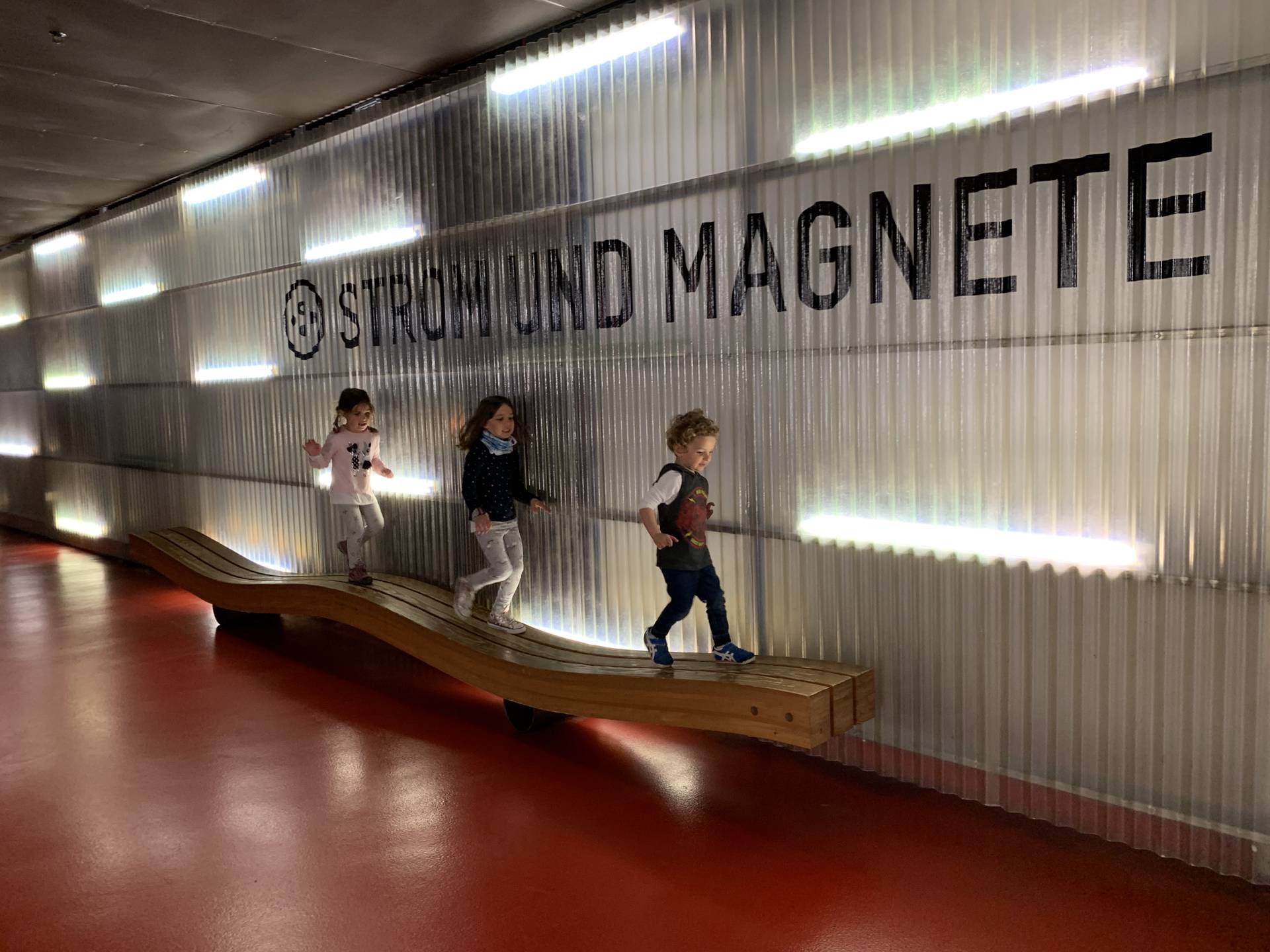 Three children walking along a wooden wavy bench in a red-floored corridor in the Swiss Science Center Technorama in Winterthur, outside the Electricity and Magnetism Section.