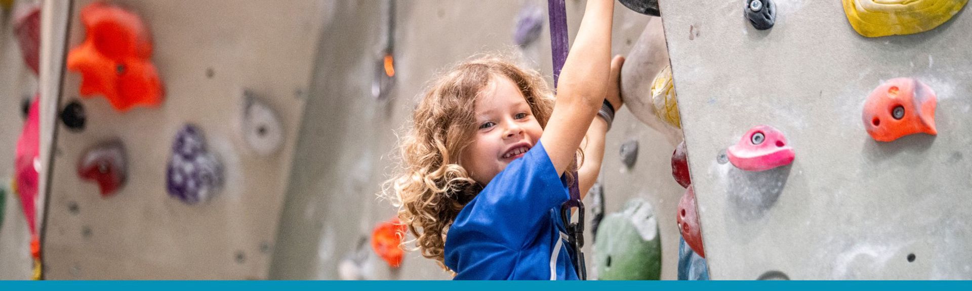 Children wearing safety harnesses climbing a colorful indoor rock wall, guided by an instructor.