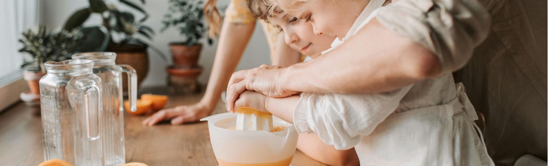 Two young children preparing fresh orange juice at a kitchen table, supported by an adult, illustrating healthy habits and family care during flu season.