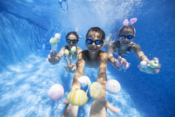 Three children are underwater inside a swimming catching colorful balls and playing games together.  They are wearing goggles and facing the camera.  