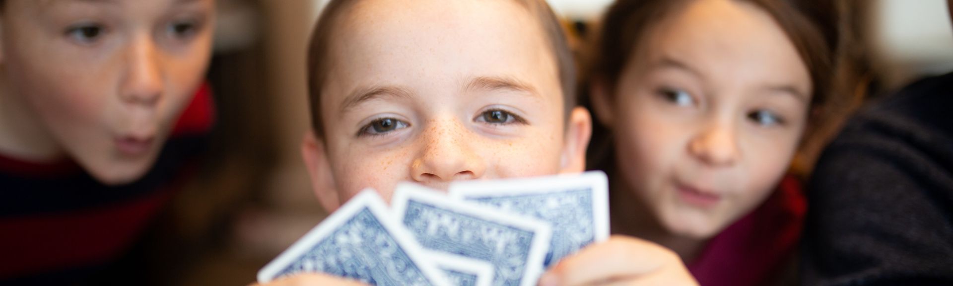 A pre-teen boy is making eye-contact with the camera while holding playing cards. Two girls behind him are peaking in his cards. It is a great pass-time activity during holidays that replaces screen time for kids, and encourages family time and social interaction.